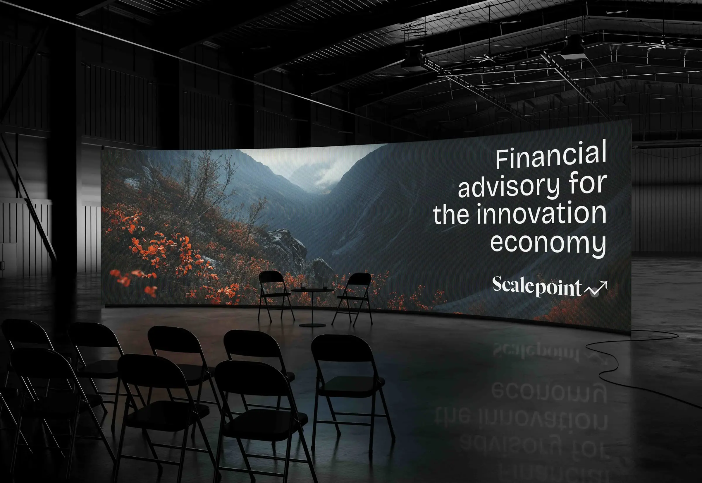 A curved LED wall in a dark warehouse reading 'Financial advisory for the innovation economy' over a mountain photograph, with folding chairs set out in front
